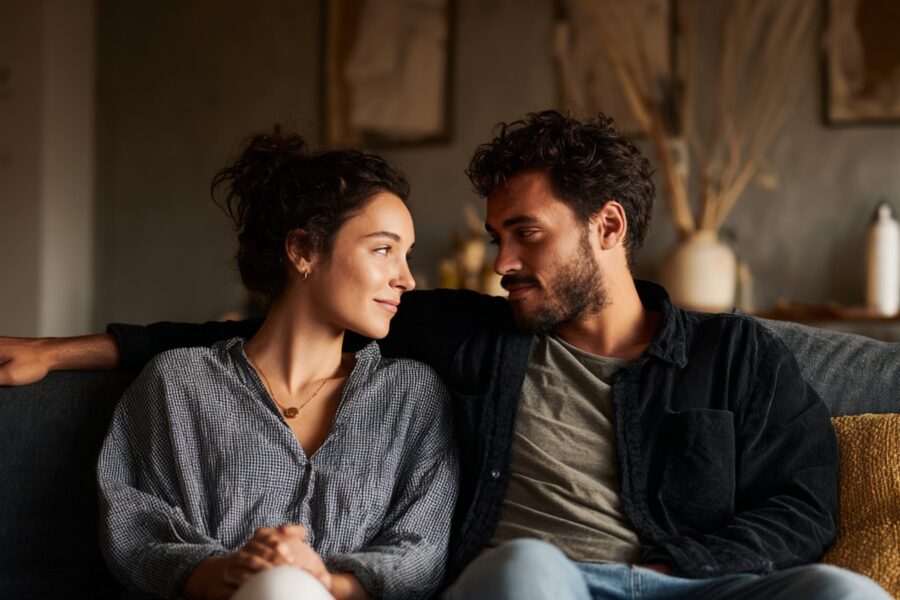 A couple sitting close together on a couch, sharing relaxed eye contact and emotional connection, representing Modern Tantra and embodied intimacy.