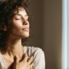 A woman stands in soft natural light with her eyes closed and hand on her chest, representing the introspective practice of tantra for singles.