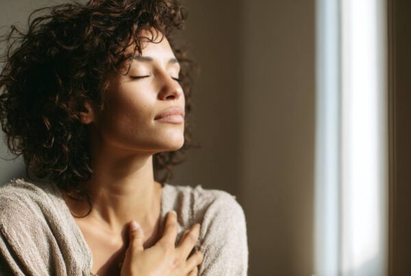 A woman stands in soft natural light with her eyes closed and hand on her chest, representing the introspective practice of tantra for singles.
