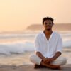 A man sits peacefully cross-legged on a beach at sunset, embodying calm presence and the reflective journey of tantra for singles.