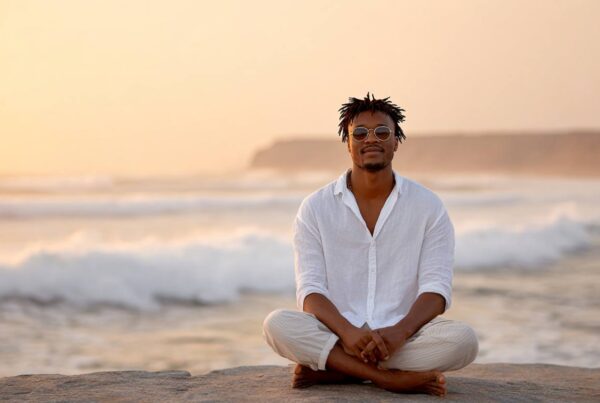 A man sits peacefully cross-legged on a beach at sunset, embodying calm presence and the reflective journey of tantra for singles.